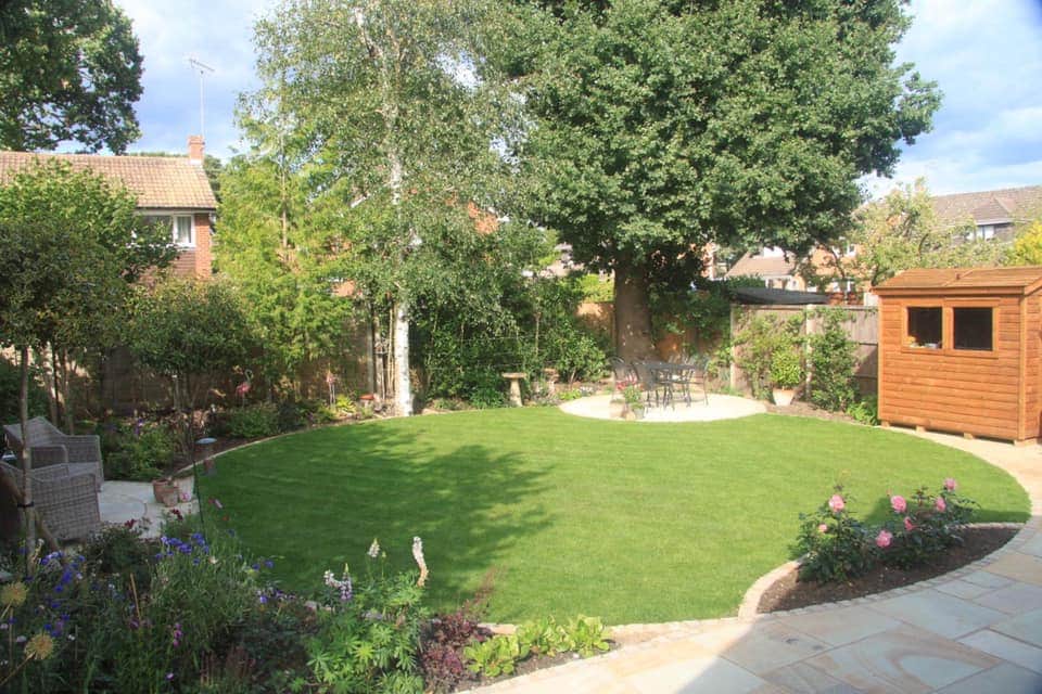 A finished back garden: a curved lawn edged in sandstone, a round seating patio under a big oak, planted borders and a shed