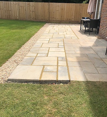 A long sandstone patio and path beside a lawn, with garden furniture at the end
