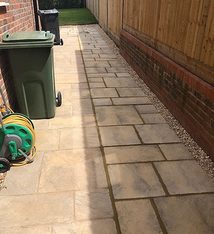A paved side passage beside a brick wall, with bins and a hose reel